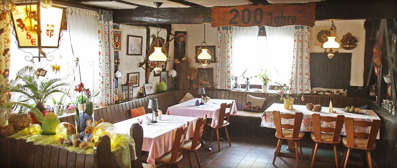 Rustikaler Gastraum mit Holzdecke, Eckbänken, gedeckten Tischen und einem Holzschild „200 Jahre“